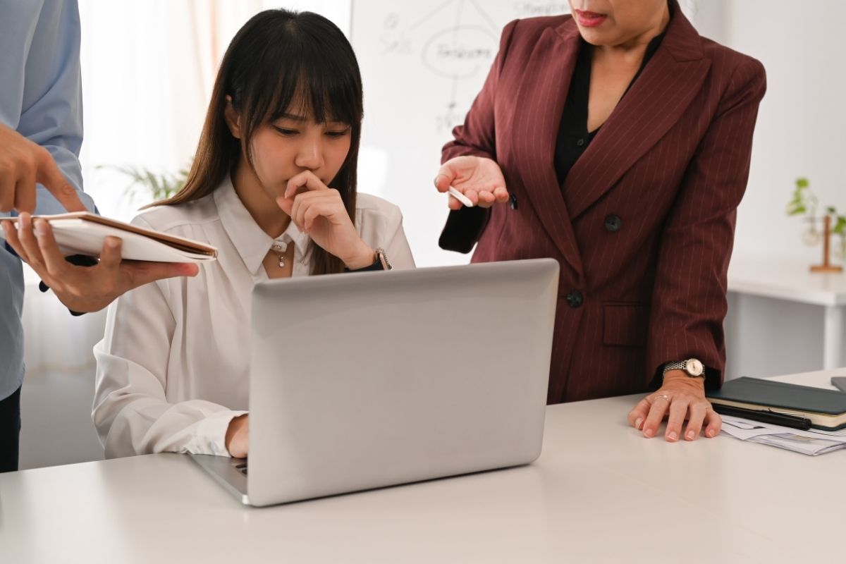 A junior communications staffer looks nervous as two more senior people look over her shoulder at her laptop