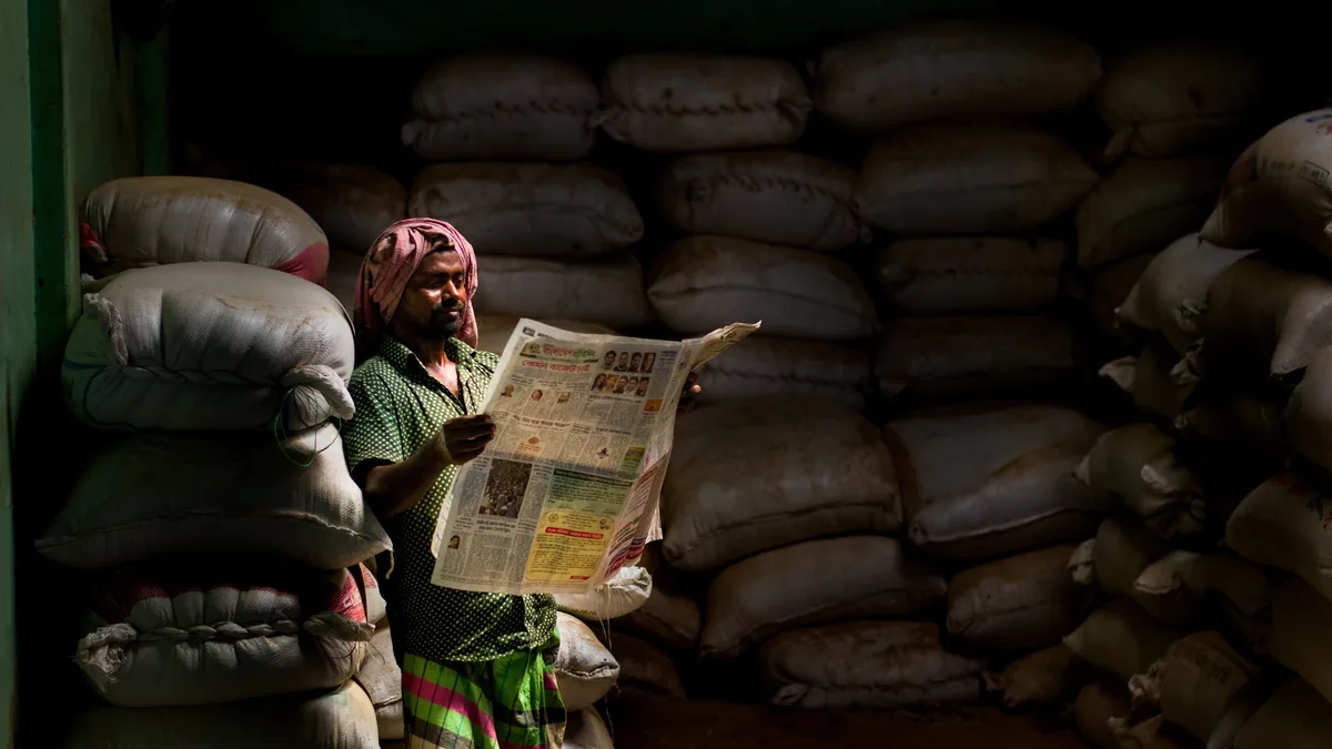 A person reads a newspaper while standing among stacked materials in a dim workspace